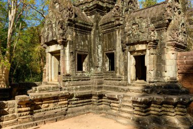 Thommanon Tapınağı 'nın antik kumtaşı cephesi, Kamboçya' nın Angkor kentindeki yüksek orman ağaçlarının arasında karmaşık oymalar ve yaşlanmış duvarlarla aydınlanıyor. Sahnede dokulu adımlar, derin gölgeler ve canlı bir görüntü yer alıyor.