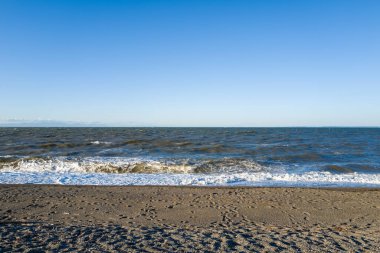 Geniş kumlu bir sahil, Ise yakınlarında bulutsuz mavi bir gökyüzünün altında dalgalarla karşılaşıyor. Ayak izleri desenli kumu aydınlatırken güneş köpüklü kıyı şeridini aydınlatarak yeni ve açık bir kıyı havası yaratıyor..