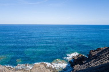 Arjantin, Sardunya yakınlarındaki engebeli, kayalık bir kıyı şeridine vuran yumuşak dalgalarla dolu derin Akdeniz manzarası. Açık gökyüzü ve güneş ışığı dingin ve açık bir atmosfer yaratır..
