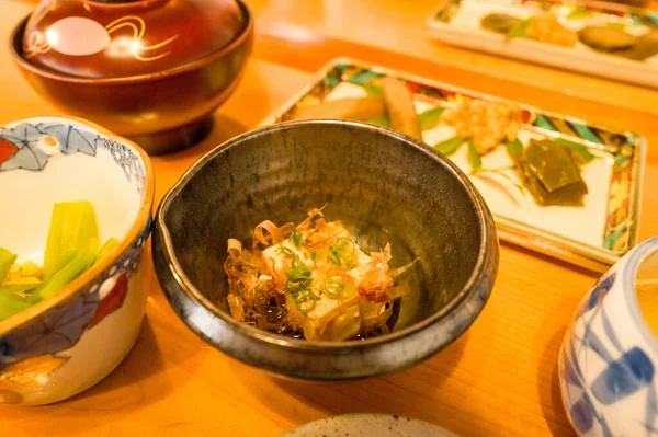 Bonito gevreği ve soğanla süslenmiş soğumuş tofu servisi, ahşap bir masada çeşitli Japon kahvaltı tabakları ile çevrili, koyu bir seramik kasede oturmaktadır. Sıcak aydınlatma geleneksel ayarların davetkar dokularını ve renklerini arttırır.