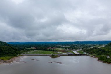 Yeşil tepeler ve alçak bulutların altında tarım alanlarıyla çevrili geniş bir rezervuar manzarası. Sahnede bir baraj yapısı, yansıtıcı su ve yemyeşil bir vadideki kırsal arazinin bir parçası yer alıyor..
