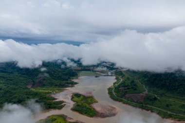 Bereketli yeşil tepelerle çevrili kıvrımlı, çamurlu bir nehrin hava perspektifi, kısmen alçak asılı bulutlarla gizlenmiş. Manzarada doğal su kanalları, yoğun bitki örtüsü ve uzak bir kırsal bölgede sisli bir atmosfer bulunur..