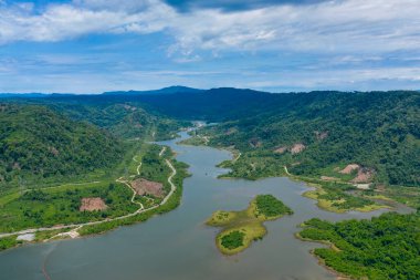 Hava perspektifi, yoğun ormanlık tepeler ve parçalı bulutlu bir gökyüzünün altındaki temiz arazi parçalarının arasında dolaşan bir nehri ortaya çıkarıyor. Manzara uzak bir kırsal bölgede dolambaçlı su, canlı yeşillik ve doğal kıvrımlara sahiptir..