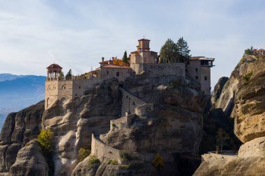 Tarihsel Varlaam Manastırı, Yunanistan 'ın Meteora kentindeki dramatik kumtaşı uçurumlarının üzerine yükseliyor. Dökümlü taş duvarlar ve yumuşak öğleden sonra ışıklarıyla aydınlanan kırmızı çatılar, dağlık bir zemin üzerine kuruluyor..
