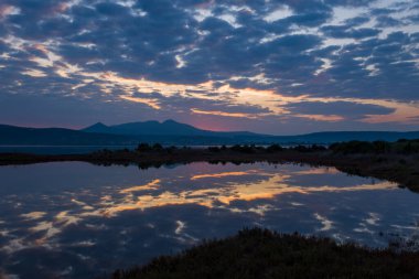 Canlı bulutlar ve dağ siluetleri, şafak vakti Yunan dağlarında bir sulak lagünün durgun yüzeyini yansıtarak, huzurlu ve karamsar bir manzara yaratıyor..