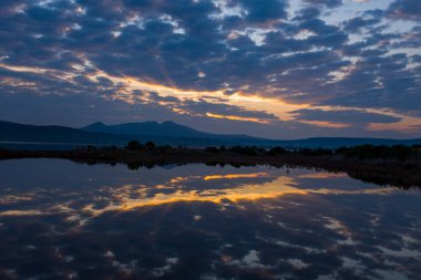 Huysuz mavi saat ışığı ve dramatik bulutlar sakin bir gölün camsı yüzeyine yansıyor. Uzak Yunan dağları, parlayan bir akşam gökyüzüne karşı siluetlenmiş..