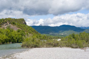Geniş çakıl taşı kıyısı, yoğun yeşil ağaçlar ve engebeli tepelerle çevrili Rio Cinca ile çarpıcı bulutlar ve uzak dağlarla kesişiyor. Parlak gün ışığı zıt dokuları ve taze atmosferi vurgular.