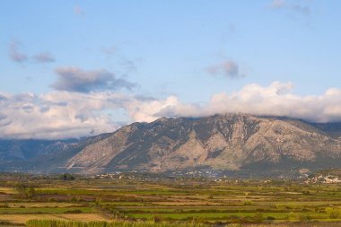 Yunanistan 'ın kırsal kesimindeki engebeli dağlara doğru uzanan katmanlı araziler, alçak bulutlar ve yumuşak öğleden sonra ışığıyla sakin bir atmosfer yaratıyor. Sahne, doğal dokuları, açık tarım arazilerini ve mavi bir yamacın altındaki uzak kayalık yamaçları ön plana çıkarıyor.