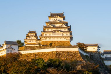Himeji Kalesi, sonbahar yaprakları ve açık mavi gökyüzü ile çevrili, altın günbatımı ışığı altında parlayan beyaz duvarları, taş surların üzerinde yüksekte duruyor..