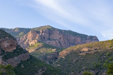 Yunanistan 'ın Kalavrita kentinin kırsal kesimindeki yeşil tepelerin üzerinde doğal mağaraları olan dik kireçtaşı kayalıklar yükseliyor. Sahnede engebeli kaya oluşumları, dağınık bitki örtüsü ve açık bir gökyüzünün altında yumuşak bir öğleden sonra ışığı yer alıyor..