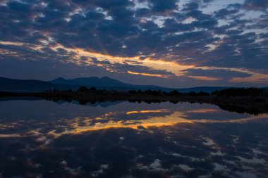 Yumuşak güneş ışınları dramatik akşam bulutlarını delip geçiyor, durgun bir lagün boyunca sıcak bir parıltı yayıyor, gökyüzünü ve Yunan dağlarındaki uzak dağ siluetini kusursuz yansıtıyor..