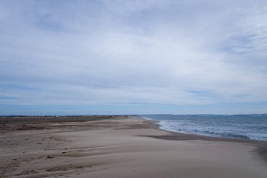Rahatsız edilmemiş geniş bir kum tabakası, Saint Louis du Rhone Limanı 'ndaki Plage Napolyon' da bulutlarla dolu yumuşak bir gökyüzünün altında hafif dalgalarla karşılaşır. Kumda ince desenler ve gün ışığını kısmak sakin bir sahil manzarası yaratır..