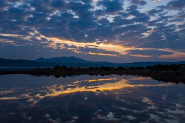 Sabahın erken saatlerinde yoğun bulutların arasından çıkan ışık, uzak mesafedeki siluetli Yunan dağları ile sakin bir sulak arazinin durgun sularına canlı yansımalar yansıtıyor..