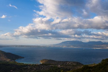 Orman tepeleri tarafından çerçevelenmiş, dinamik, güneşli bulutlarla dolu bir gökyüzünün altındaki uzak sıradağlarla dolu derin mavi bir koy üzerinde geniş bir manzara. Sahne, sükunet ve doğal ihtişam saçıyor..