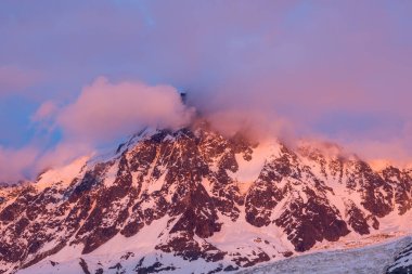 Yumuşak altın ve pembe ışık Aiguille du Midi 'nin engebeli, karlı uçurumlarını aydınlatıyor. Sisli bulutlar Fransız Alpleri' nin dramatik alp yüzeyinde sürükleniyor..