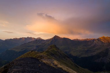 Yumuşak akşam ışığı, Pireneler 'deki çimenli bir dağ sırtında pastel bir parıltı yayar, dramatik tepeler ve yumuşak bulutlar, sakin, atmosferik bir manzara yaratır..
