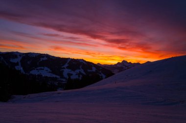 Dramatik kış günbatımı, kar kaplı kayak yamaçlarının ve Megeve 'nin siluetli dağlarının üzerinde gökyüzünü canlı turuncu ve mor renklerle boyar. Huzurlu dağ manzarası renkli alacakaranlıkla yıkanır..