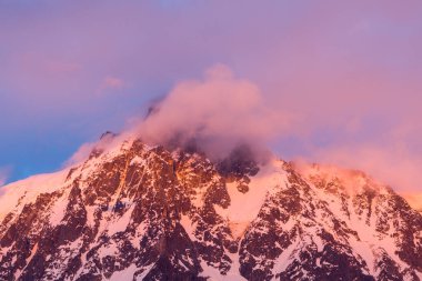 Sıcak pembe ve turuncu bulutlar Aiguille du Midi 'nin engebeli, karla kaplı zirvesinde sürükleniyor günbatımının son ışınlarını yakalıyor ve dramatik alp dokusunu vurguluyor..