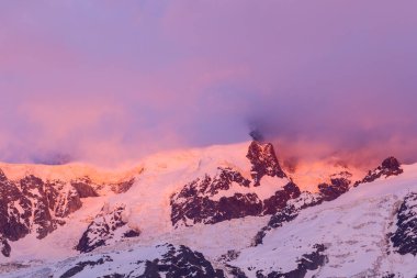 Parlak pembe ve turuncu gün doğumu ışığı karla kaplı Mont Blanc du Tacul tepelerini yıkıyor, engebeli dağların üzerinde dramatik bulutlar dönüyor..