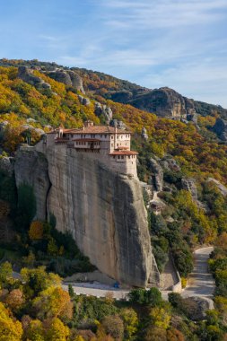 Tarihsel Roussanou Manastırı, Yunanistan 'ın Meteora kentinde canlı sonbahar yapraklarıyla çevrili dik bir kumtaşı sütunun üzerinde çarpıcı bir şekilde duruyor. Sıcak güneş ışığı manzaranın doğal dokularını ve toprak tonlarını güçlendirir..