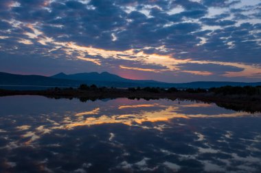 Bulutla dolu gündoğumu gökyüzü sakin sulak bir göletin sakin yüzeyine yansıyor, ufukta uzak Yunan dağlarının silueti ve yumuşak bir sabah ışığı barışçıl bir hava yaratıyor..
