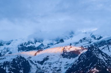 Sıcak güneş ışığı karla kaplı Mont Blanc du Tacul 'un dramatik mavi gölgelere ve yüksek dağlık arazide dönen bulutlara karşı bir bölümünü kısaca aydınlatır..