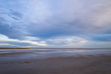Yumuşak mavi ve lavanta tonları ile boyanmış dramatik bir gökyüzünün altında yansıtıcı ıslak kumlar ufka doğru uzanır. İnce dokular ve sakin ışık barışçıl, minimalist bir kıyı havasını çağrıştırıyor..