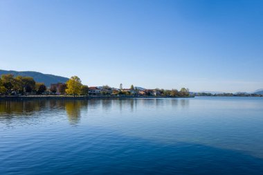 Durgun su, uzak tepeler ve berrak mavi gökyüzünün sakin bir atmosfer yarattığı Ioannina Gölü kıyısındaki göl kenarı ve sonbahar ağaçlarını yansıtır. Sahnede yumuşak doğal ışık, hafif dalgalar ve huzurlu bir şehir var.