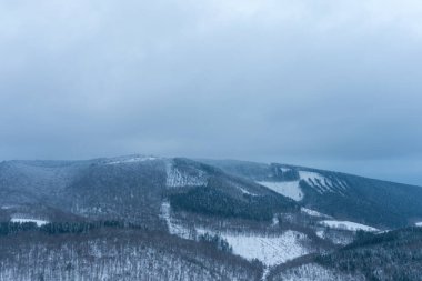 Morvan bölgesinde karla kaplı dağ sırtı ve yoğun kış ormanı bulutlu bir gökyüzü altında geniş bir manzara. Sessiz mavi tonlar ve katmanlı yamaçlar sessiz ve uzak bir atmosferi çağrıştırıyor..