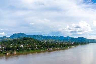 Mekong Nehri 'nin yemyeşil tepeler ve Laos' un Luang Prabang kasabasının yanında akan geniş manzarası. Dağılmış bulutlu mavi gökyüzü ormanlık dağların üzerine yayılarak sakin bir tropikal manzara yaratıyor..