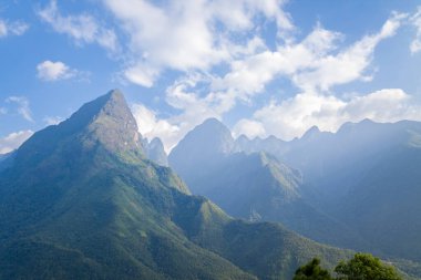 Fansipan 'ın sarp yeşil dağları parlak mavi bir gökyüzünün altında yükseliyor. Yumuşak güneş ışığı engebeli yamaçları aydınlatıyor ve dağınık bulutlar havada sürükleniyor..