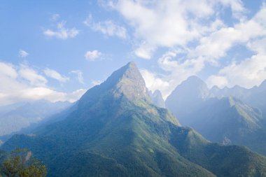 Kuzey Vietnam 'ın Fansipan bölgesindeki yamaçları kaplayan sisli tepeler ve yemyeşil ormanlarla parçalı bulutlu bir gökyüzünün altında dramatik bir yeşil dağ keskin bir şekilde yükseliyor..