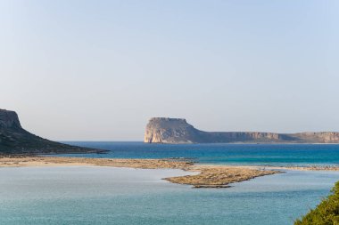 Girit 'in Balos kenti yakınlarındaki çarpıcı düzlüklerdeki bir adaya doğru sakin bir kum tabakası sığ mavi lagün suları boyunca kıvrılır. Manzara, solgun ve bulutsuz bir gökyüzünün altında güneşli ve geniştir..