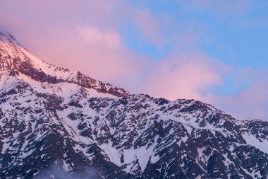 Hafif pembe ve mavi bulutlar Aiguille de Bionnassay 'in engebeli, karlı yamaçları üzerinde sürükleniyor. Yumuşak bir sabah ışığı Alp sırtında huzur dolu bir parıltı bırakıyor..