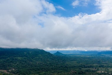 Yoğun yeşil tepelerin ve ormanların geniş manzarası yoğun, sürüklenen bulutların ve mavi gökyüzünün altında uzanır. Manzara, Laos kırsalının tipik rutubetli atmosferi ve katmanlı bitki örtüsünü vurgular..