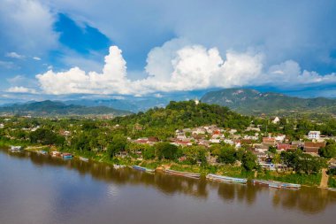 Luang Prabang, Laos 'un yüksek açılı manzarası. Mekong Nehri' nin yanındaki yoğun yeşilliklerin arasında geleneksel çatıları var. Dramatik kümülüs bulutları orman tepeleri ve uzak mavi dağlar üzerinde yükselir canlı bir gökyüzünün altında.