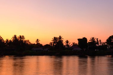 Güney Laos 'un 4000 ada bölgesindeki Mekong Nehri boyunca uzanan palmiye ağaçları ve geleneksel nehir kenarındaki evler ılık akşam ışığı altında. Durgun su, gökyüzünün turuncu ve pembe renklerini yansıtarak sakin bir tropikal manzara yaratır..