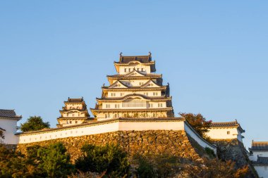 İkonik beyaz Himeji Kalesi taştan duvarların ve sonbahar yapraklarının üzerinde yükseliyor, açık mavi gökyüzüne karşı ılık bir akşam güneşi ışığında parlıyor..