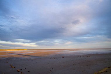 Altın ışık, yumuşak mavi ve lavanta bulutlarıyla dolu bir gökyüzünün altında, Camargue sahilinin geniş ve boş gelgit düzlüklerini nazikçe aydınlatarak, huzurlu ve geniş bir kıyı manzarası yaratıyor..
