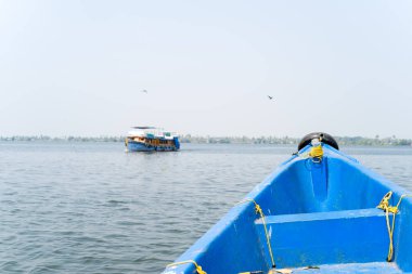 Canlı mavi bir kanonun pruvası Hindistan 'ın Alappuzha kentindeki uzak bir tekneyi işaret ediyor. Sakin bir su ve puslu bir gökyüzü rahatlatıcı, yolculuk hissi veren açık bir atmosfer yaratır..