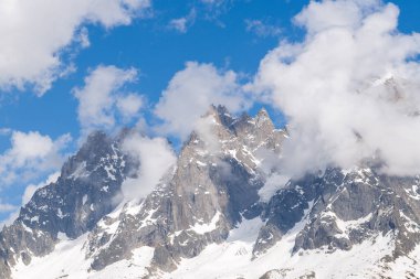 Keskin, engebeli dağ zirveleri kısmen dönen bulutlar tarafından gizleniyor. Fransız Alpleri 'ndeki canlı mavi gökyüzünün altındaki sarp kayalara yapışmış kar parçalarıyla..