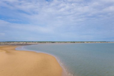 Geniş bir kumsalın kıvrımlı kıyı şeridi yumuşak bulutlu bir gökyüzünün altında sakin Atlantik sularıyla buluşuyor. Uzak alçak binalar ve yeşil kuşak Ile de Re yakınlarında sakin bir kıyı atmosferini çağrıştırıyor..