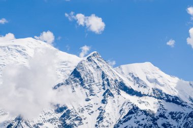 Aiguille de Bionnassay 'in karla kaplı zirvesi Mont Blanc' ın arkaplanına karşı duruyor. Parlak mavi gökyüzünün altında canlı dokular ve dağınık bulutlar..