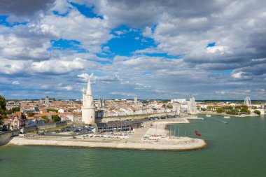 La Rochelles 'in ikonik ortaçağ kuleleri, taş duvarları ve Atlantik kıyısı boyunca turuncu kiremitli çatılardan oluşan yoğun bir kasabası olan tarihi limanının geniş hava perspektifi. Sahnede sakin yeşil bir su, bir marina ve dramatik bir gökyüzü yer alıyor.