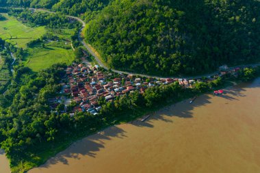 Yoğun yeşil tepeler ve Mekong nehrinin çamurlu suları arasında yer alan nehir kenarındaki küçük bir köyün hava manzarası. Sahnede kırmızı çatılı evler, yemyeşil bitki örtüsü ve Güneydoğu Asya 'daki parlak güneş ışığı altında ekilmiş tarlalar yer alıyor..