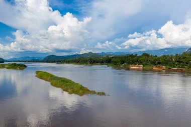 Büyük kümülüs bulutları Mekong Nehri 'nin yukarısında, Luang Prabang, Laos yakınlarındaki çimenli adaları ve tropikal kıyıları geçerken. Ahşap botlar bereketli kıyı şeridi boyunca uzanır, uzak dağlar parlak gün ışığında manzaralı bir zemin oluşturur..
