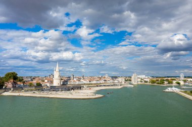 La Rochelles tarihi limanının geniş manzarası ikonik taş kuleler, rıhtım binaları ve desenli bulutlarla dolu dramatik bir gökyüzü. Sahnede sakin yeşil su, ortaçağ mimarisi ve Atlantik kıyısı boyunca canlı eski bir kasaba yer alıyor..