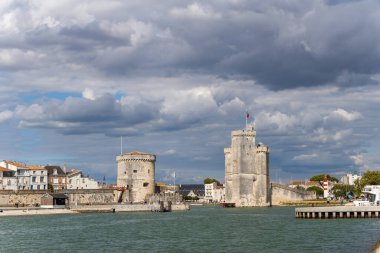 İkonik ortaçağ taş kuleleri dramatik bir gökyüzünün altındaki tarihi La Rochelle limanının girişini koruyor. Sahnede güçlendirilmiş mimari, rıhtım binaları ve Atlantik kıyısı boyunca dokulu bulutlar yer alıyor..