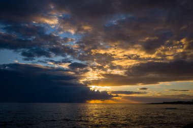 Altın ışınlar Adriyatik Denizi üzerindeki dramatik bulut katmanlarını delip, alacakaranlıkta sıcak yansımalarla suyu aydınlatıyor. Sahne, karanlık bulutlar ve sakin deniz manzarası boyunca parlayan gökyüzü arasında çarpıcı bir kontrastı gösteriyor..