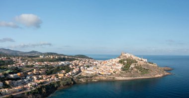Castelsardo 'nun geniş hava manzarası kayalık bir çıkıntıya tünemiş, pastel binalar sakin Akdeniz' e dökülüyor ve arka planda açık mavi bir gökyüzünün altında yuvarlanan tepeler var..